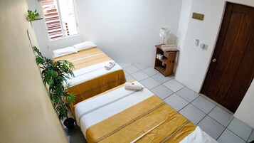 A hotel room with three beds, a small wooden shelf with towels, a potted plant, and a door.