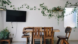 A dining area with a wooden table and chairs, a television mounted on the wall, and green vines decorating the space. - Brisamar Guesthouse (Puerto Escondido)