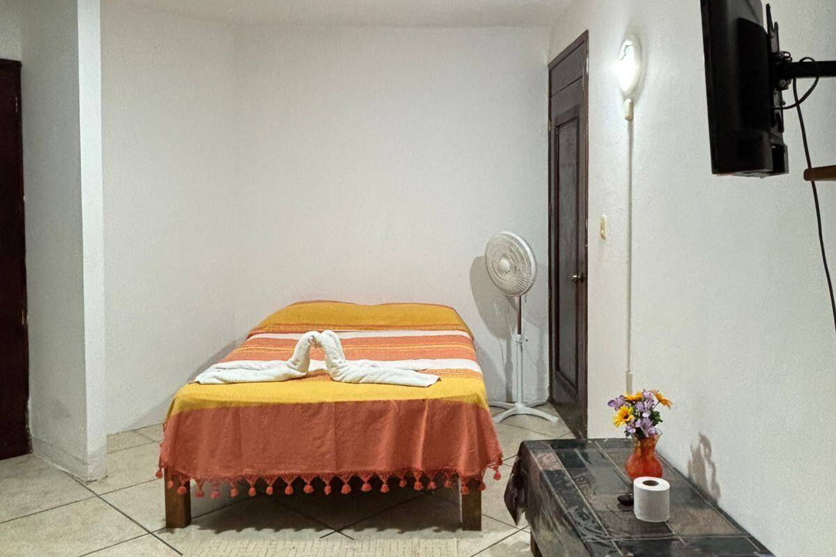 Ein kleiner, ordentlicher Raum mit einem Bett, einem Ventilator, einem Fernseher und einem kleinen Tisch mit einer Blumenvase.