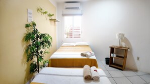 A hotel room with two beds, a window with blinds, a wall-mounted air conditioner, a small table with a lamp, and a potted plant. - Brisamar Guesthouse (Puerto Escondido)