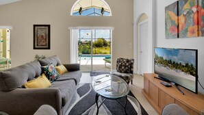 Ein Wohnzimmer mit einem grauen Sofa, einem Glastisch, einem Fernseher auf einem Holzständer und einem Papageienbild an der Wand.