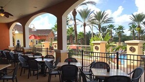 An outdoor dining area with tables and chairs, surrounded by a fence, with palm trees and a pool in the background. - RP144BD - Disney Regal Palms Townhome near Disney - Come on by! (Davenport)