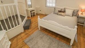 Une chambre à coucher avec un lit blanc, un bureau, une chaise, un petit banc et une fenêtre avec des stores.