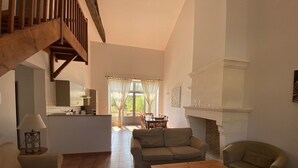 A spacious living room with a wooden staircase, a fireplace, and a dining area visible through a window. - Les Houx - Old barn with private swimming pool (Chalais)