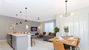 Eine moderne Küche mit Kochinsel, Essbereich mit Holztisch und Stühlen sowie ein Wohnzimmer mit Sofa und Fernseher.