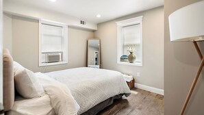 Un dormitorio con cama, dos ventanas con persianas, un espejo y una lámpara de pie.
