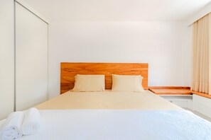 A neatly made bed with a wooden headboard, a white wall, and a window with curtains.