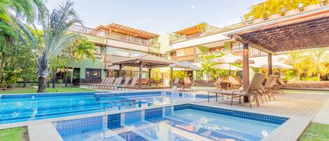 A swimming pool with lounge chairs and umbrellas, surrounded by greenery and a multi-story building with balconies.
