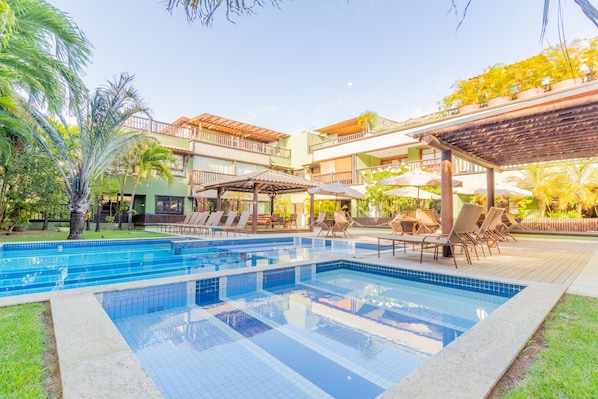 A swimming pool with lounge chairs and umbrellas, surrounded by greenery and a multi-story building with balconies.