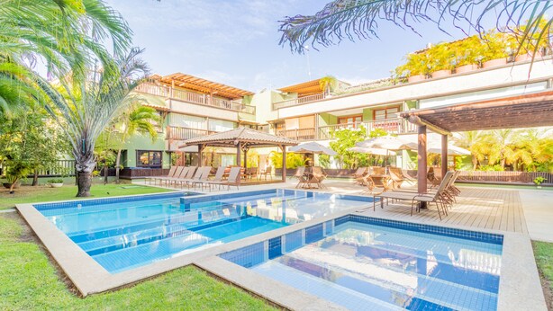A swimming pool with lounge chairs and umbrellas, surrounded by greenery and a multi-story building. - PF36 Ótimo Quarto e Sala a 25m da Praia (Mata de São João)