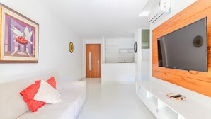 A modern living room with a white sofa, a flat-screen TV mounted on a wooden panel, and a kitchen in the background. - PF36 Ótimo Quarto e Sala a 25m da Praia (Mata de São João)