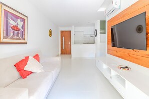 A modern living room with a white sofa, a flat-screen TV mounted on a wooden panel, and a kitchen in the background.
