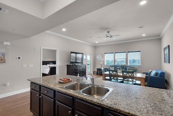 A modern kitchen with granite countertops, stainless steel sink, and dark cabinetry. There is a dining area with a wooden table and blue chairs.