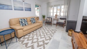 A living room with a beige sofa, patterned cushions, a blue side table, a patterned rug, a wooden sideboard, and a television. - Los Flamencos apto N56 (Manilva)