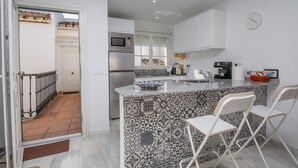 A modern kitchen with white cabinets, a stainless steel refrigerator, a microwave, and a coffee machine. There is a patterned backsplash and a small island with two white chairs. - Los Flamencos apto N56 (Manilva)