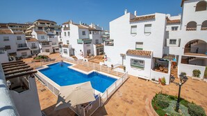 A rooftop swimming pool surrounded by white buildings with terracotta roofs, a clear sky, and a small garden area. - Los Flamencos apto N56 (Manilva)