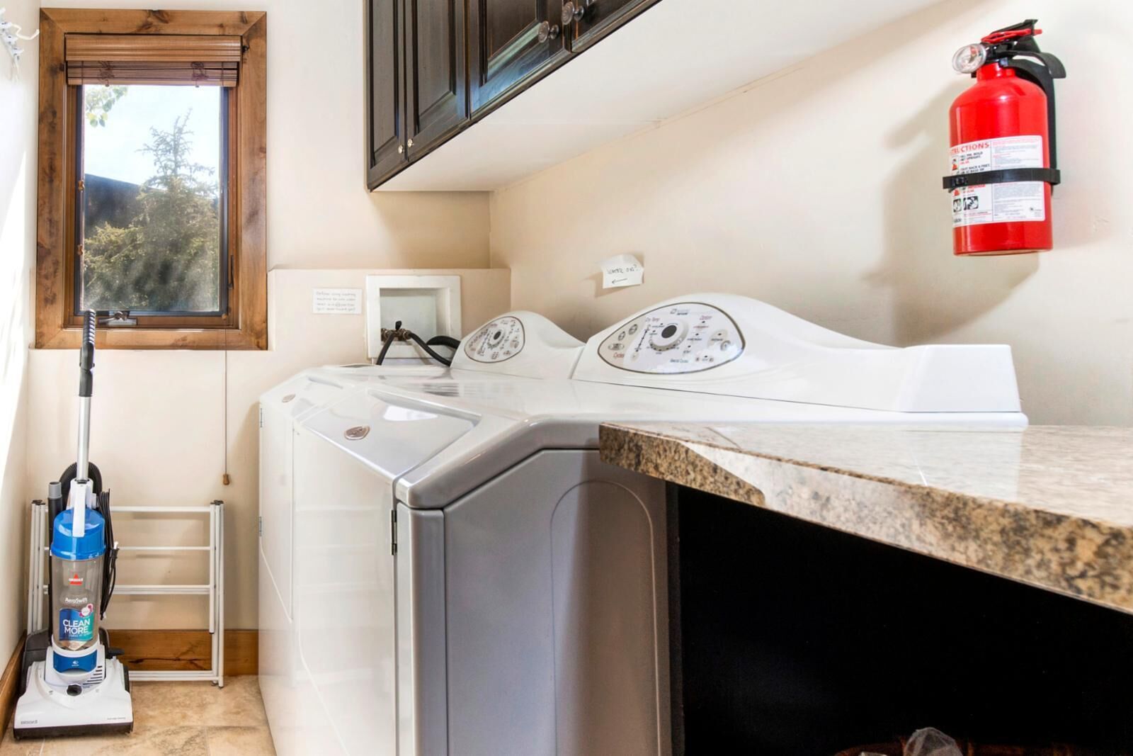 A laundry area with a front-loading washer and dryer, a vacuum cleaner, a fire extinguisher, and a window with a view of trees.