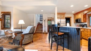 Un vaste espace de vie comprenant un canapé, des fauteuils, une table basse, une lampe, ainsi qu’une cuisine dotée de meubles en bois et d’un coin bar.