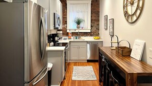Een functionele keuken met witte kasten, een roestvrijstalen koelkast, een magnetron en een spoelbak. Er staat een houten eettafel met stoelen en een decoratieve klok hangt aan de muur.