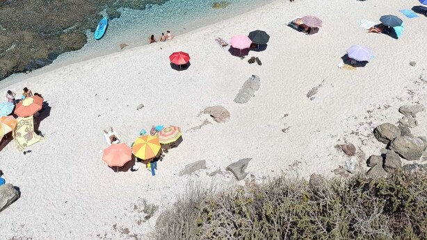 Une plage à l’eau turquoise et limpide, parsemée de rochers et où des gens se prélassent au soleil sous des parasols colorés.