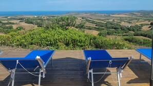Une terrasse agrémentée de deux fauteuils bleus et de parasols blancs, offrant une vue panoramique sur un paysage pittoresque, avec des vignobles et une vue lointaine sur la mer.