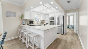 A modern kitchen with white cabinetry, a central island with bar stools, stainless steel appliances, and a 'HAPPY PLACE' sign on the wall. - Cozy Family Beach Home, Golf Cart and near  StAndrews PCB (Panama City)