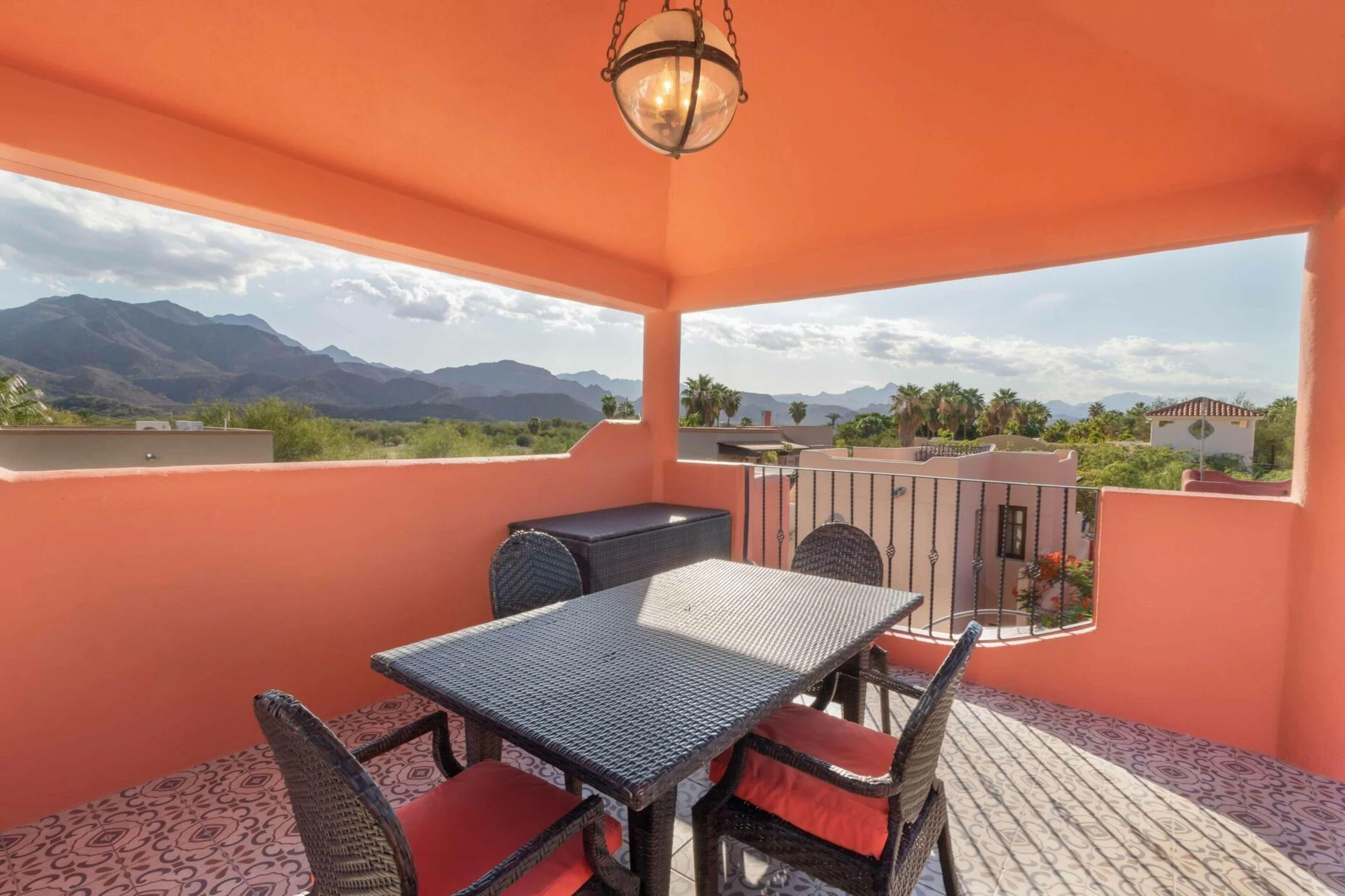 Un balcon avec une table et des chaises, un luminaire suspendu, et une vue sur les montagnes et la verdure.