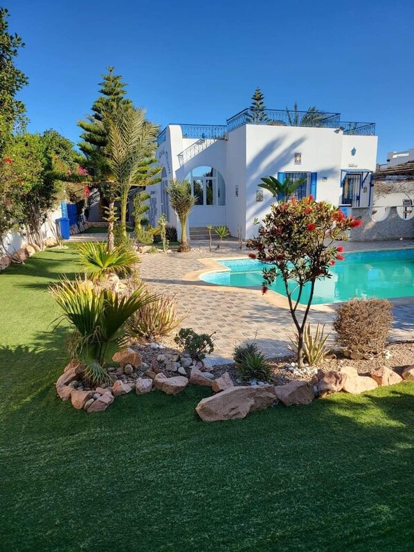 A modern house with a swimming pool, surrounded by a well-maintained garden with various plants and a stone pathway.