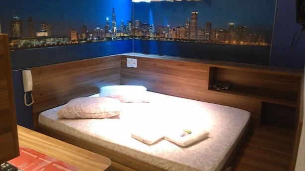 A room with a bed, a wooden headboard, a nightstand, and a cityscape mural on the wall. - Hotel Namorata Expo Inn (São Paulo)