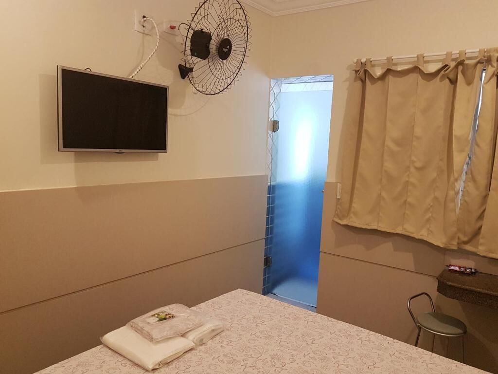 Une chambre d’hôtel avec un lit, un téléviseur fixé au mur, un ventilateur, une fenêtre avec des rideaux et un petit tabouret.