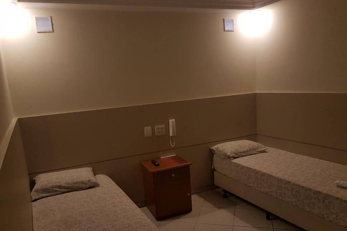 Una habitación pequeña y con poca luz, con dos camas individuales, una mesita de noche de madera y un teléfono fijo en la pared.