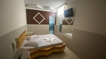 Quarto de hotel moderno com uma cama, mesa de cabeceira, televisão e banheiro visível por uma porta de vidro.