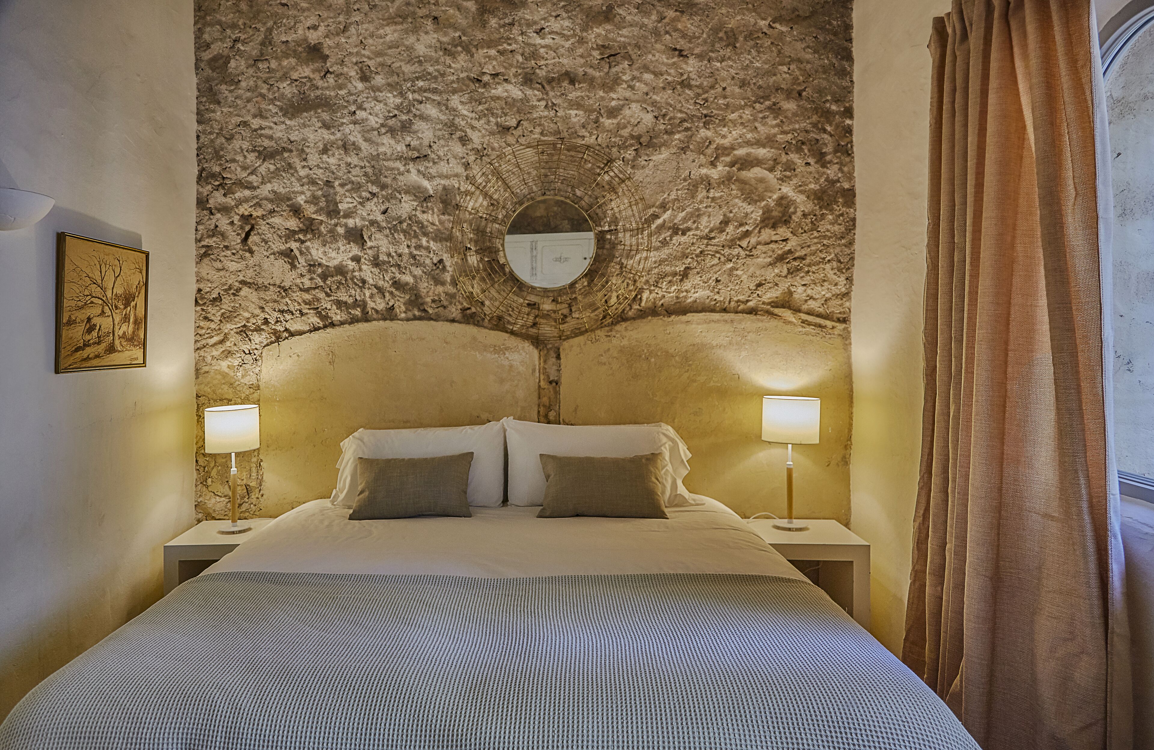 Un dormitorio con pared de piedra, un espejo redondo, dos mesitas de noche con lámparas y una cama con almohadas.