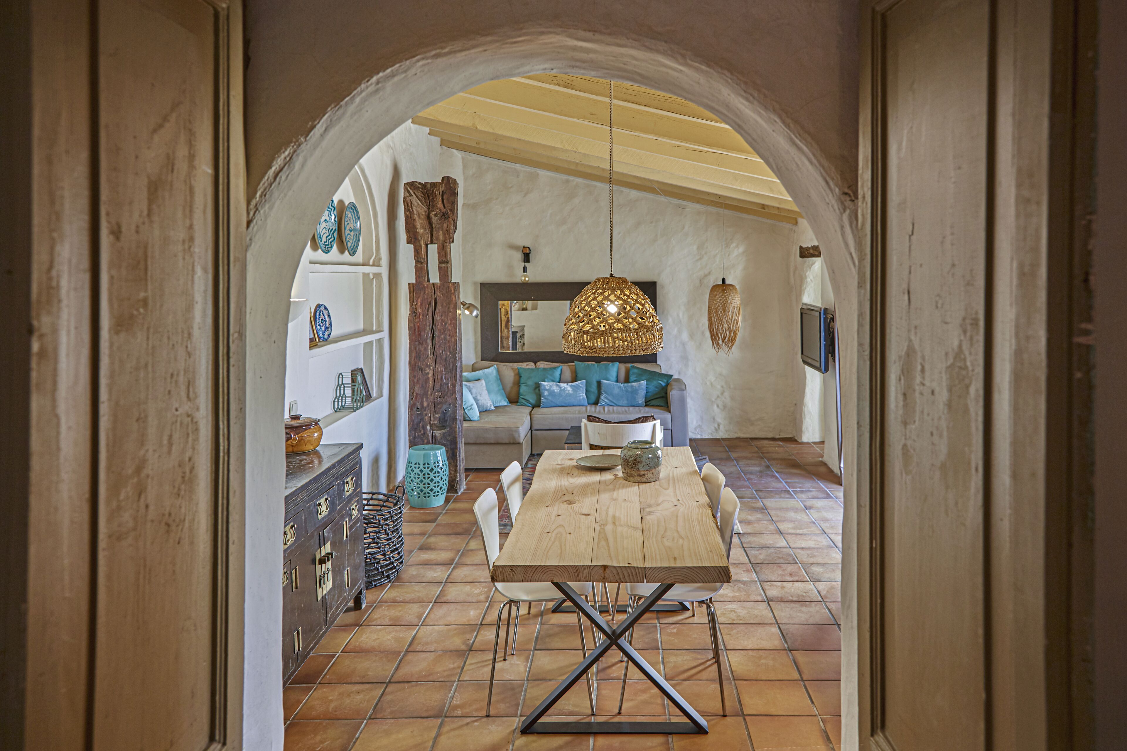 A dining area with a wooden table and chairs, a hanging light fixture, and a sofa with cushions.