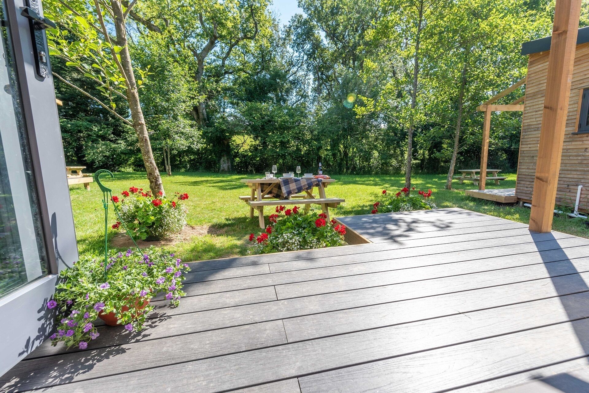 A wooden deck with a picnic table set up in a garden with a wooden fence and trees.