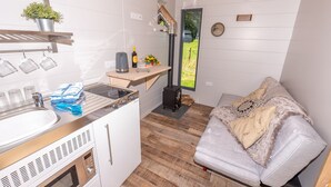 Cocina compacta con estufa, pileta y tazas colgando. Hay una mesita con una botella y una planta. Un sofá con una manta está en un rincón, y una ventana muestra una vista verde al exterior.