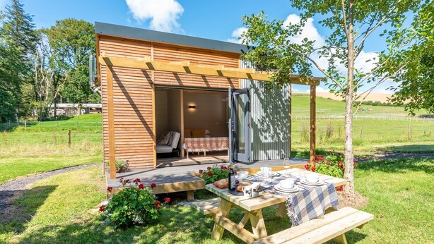 Una cabaña de madera con un área de estar al aire libre techada, una mesa de picnic y un pequeño jardín.