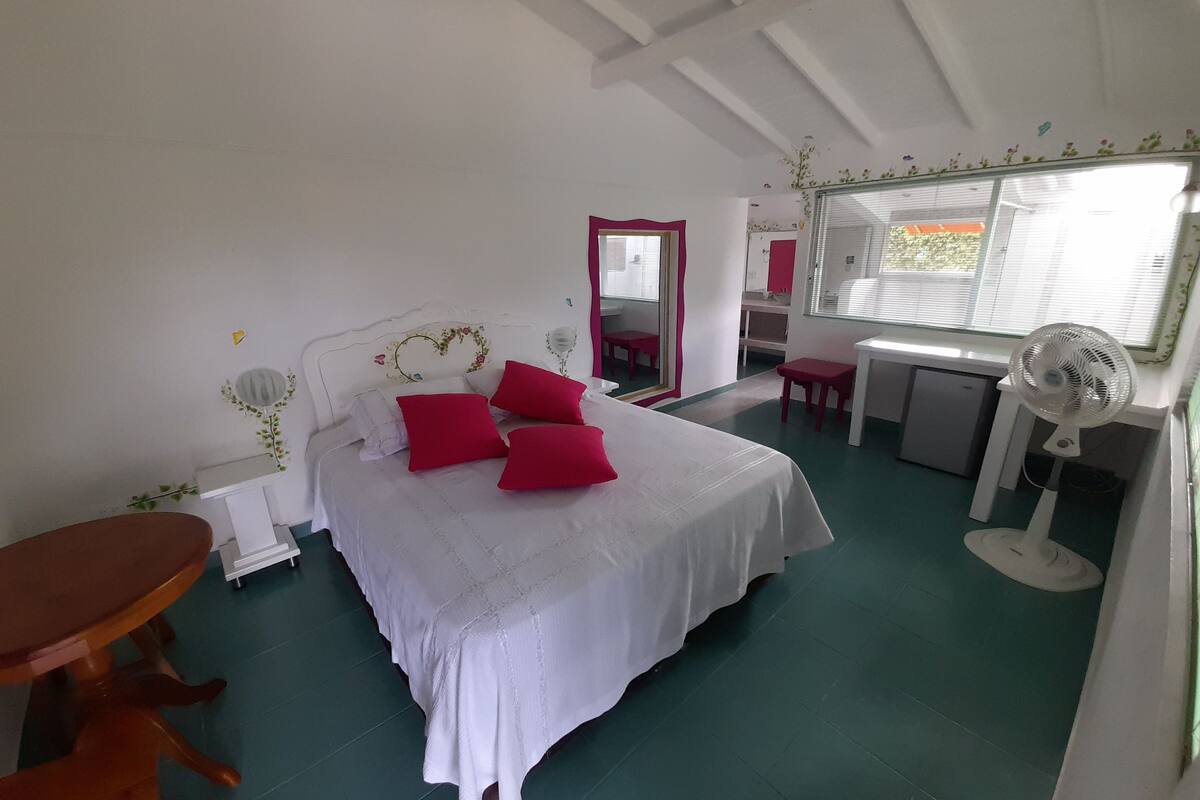 A bedroom with a bed, a desk, a chair, a table, and a mirror.