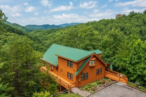 A wooden cabin with a green roof, surrounded by dense forest and hills.