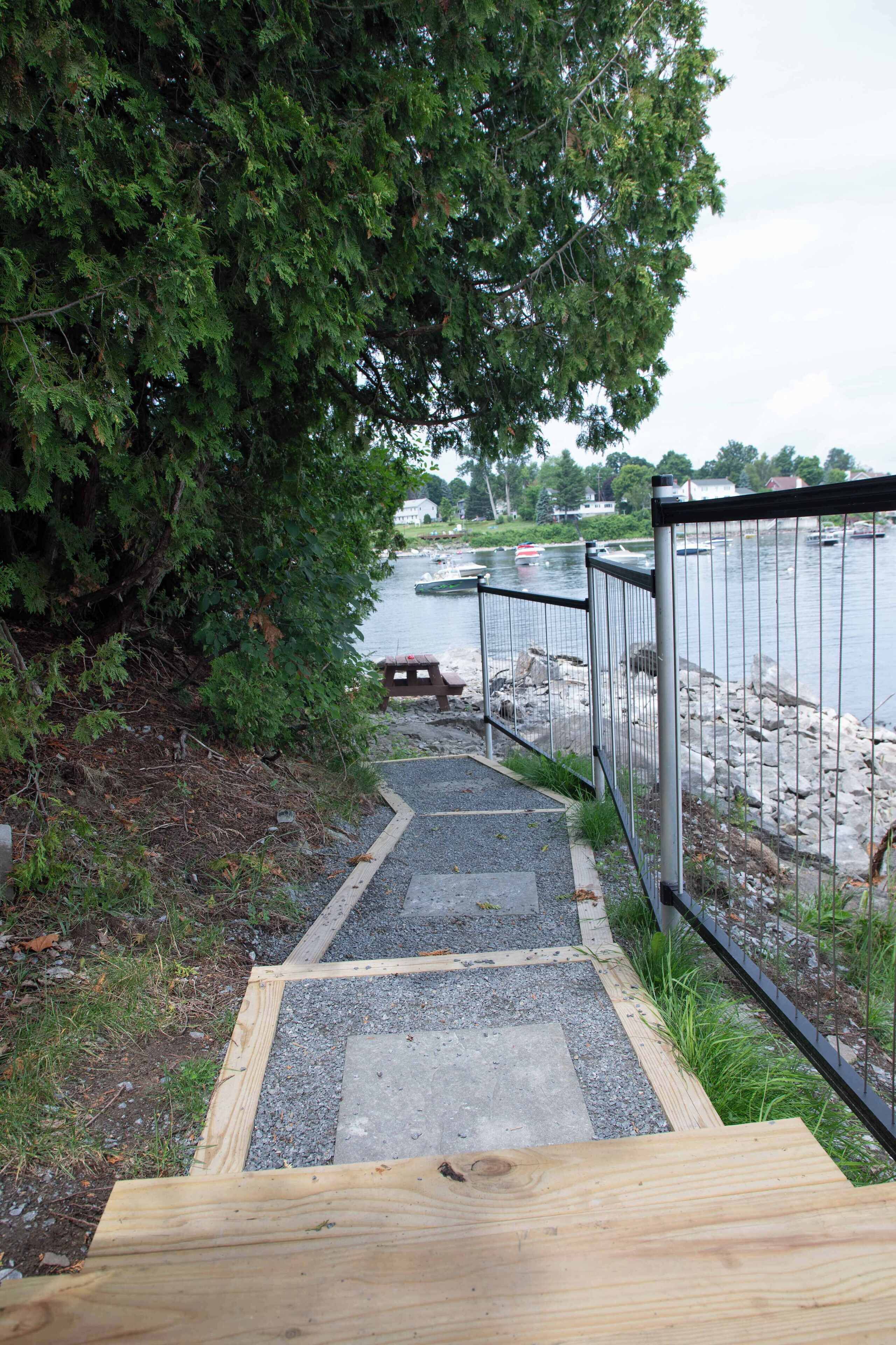 Un sendero de madera que conduce a una zona frente al agua, con una valla metálica y un banco.