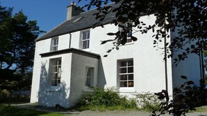 A white two-story house with a chimney, a front porch, and a small garden area. - Tullochard House (Dunvegan)