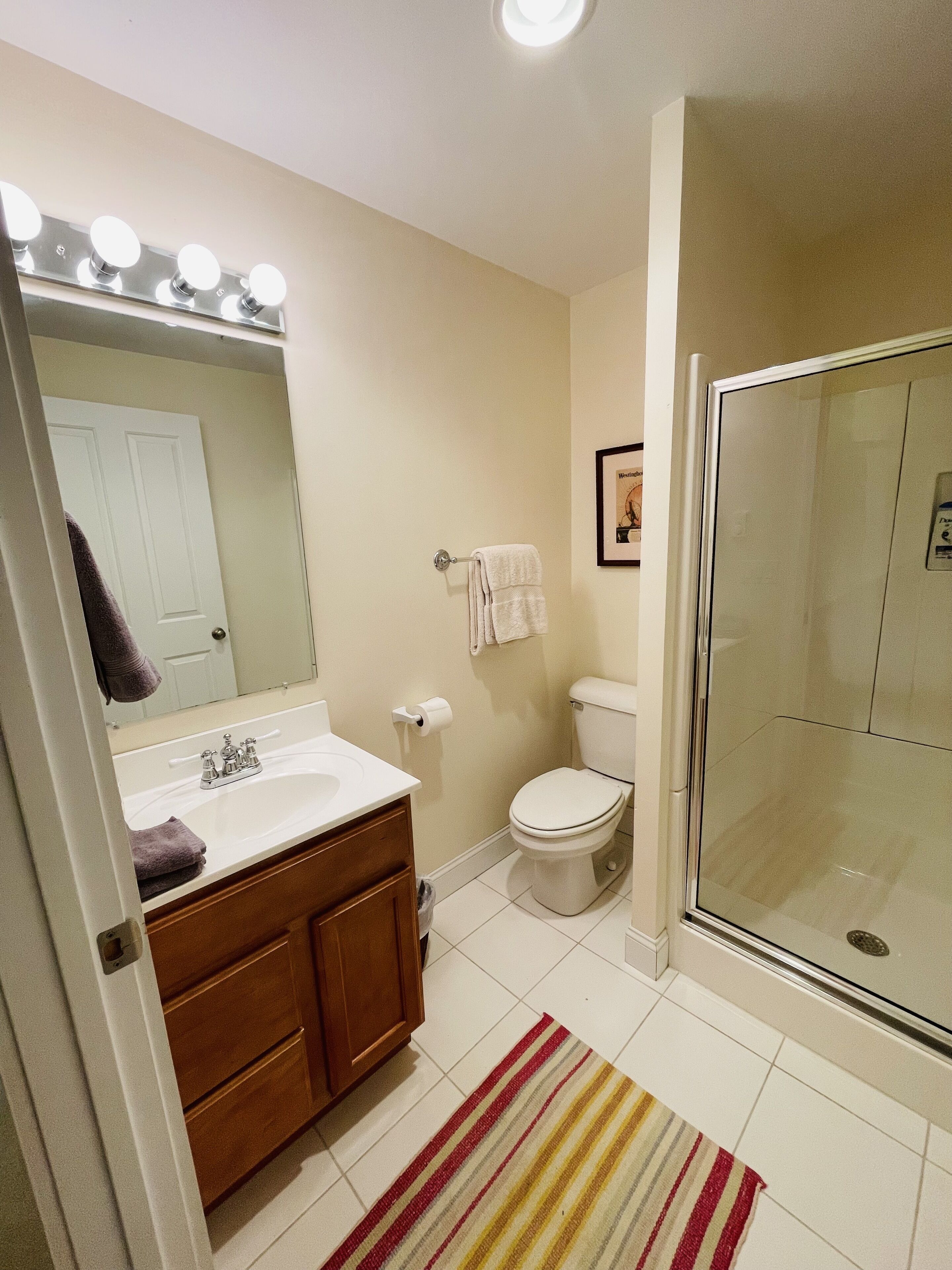 A bathroom with a white sink, mirror, and toilet. There is a shower area with a glass door and a striped rug on the floor.