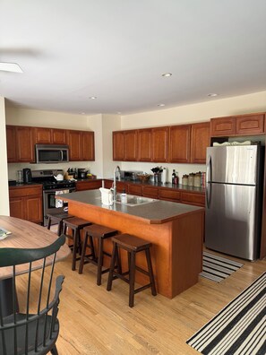 A modern kitchen with wooden cabinets, stainless steel appliances, a central island with stools, and a dining area with a table and chairs. - Perfect location in the heart of downtown Lenox (Lenox)
