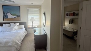 A bedroom with a bed, a desk, a chair, a lamp, a painting, and a bathroom visible through an open door. - Perfect location in the heart of downtown Lenox (Lenox)