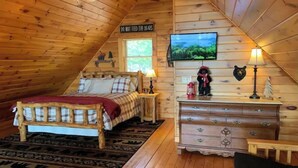 Un dormitorio acogedor en el ático, con techo inclinado de madera, una cama con colcha de cuadros, un aparador con cajones, un televisor y una decoración temática de osos.