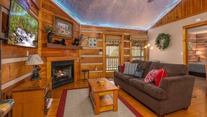Interior acolhedor de uma cabana com lareira, paredes de madeira, um sofá e uma TV fixada na parede.