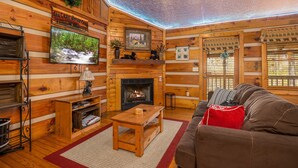 Interior acolhedor de uma cabana com lareira, paredes de madeira, uma televisão e um sofá.