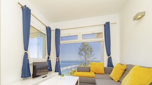 Un salon moderne avec un canapé, un téléviseur et une table. De grandes fenêtres sont ornées de rideaux, et l’on peut admirer la vue sur la mer. Une plante en pot complète l’ensemble.