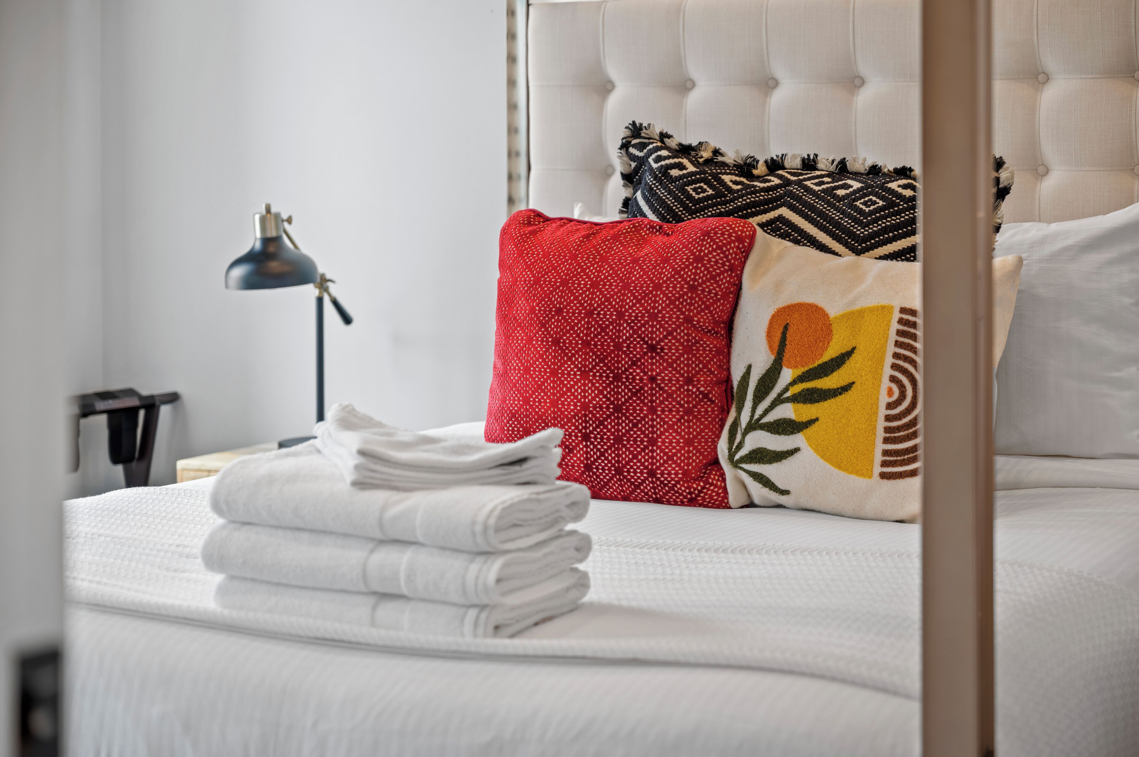 A neatly made bed with white linens, a red patterned pillow, and a decorative pillow with a sun design.
