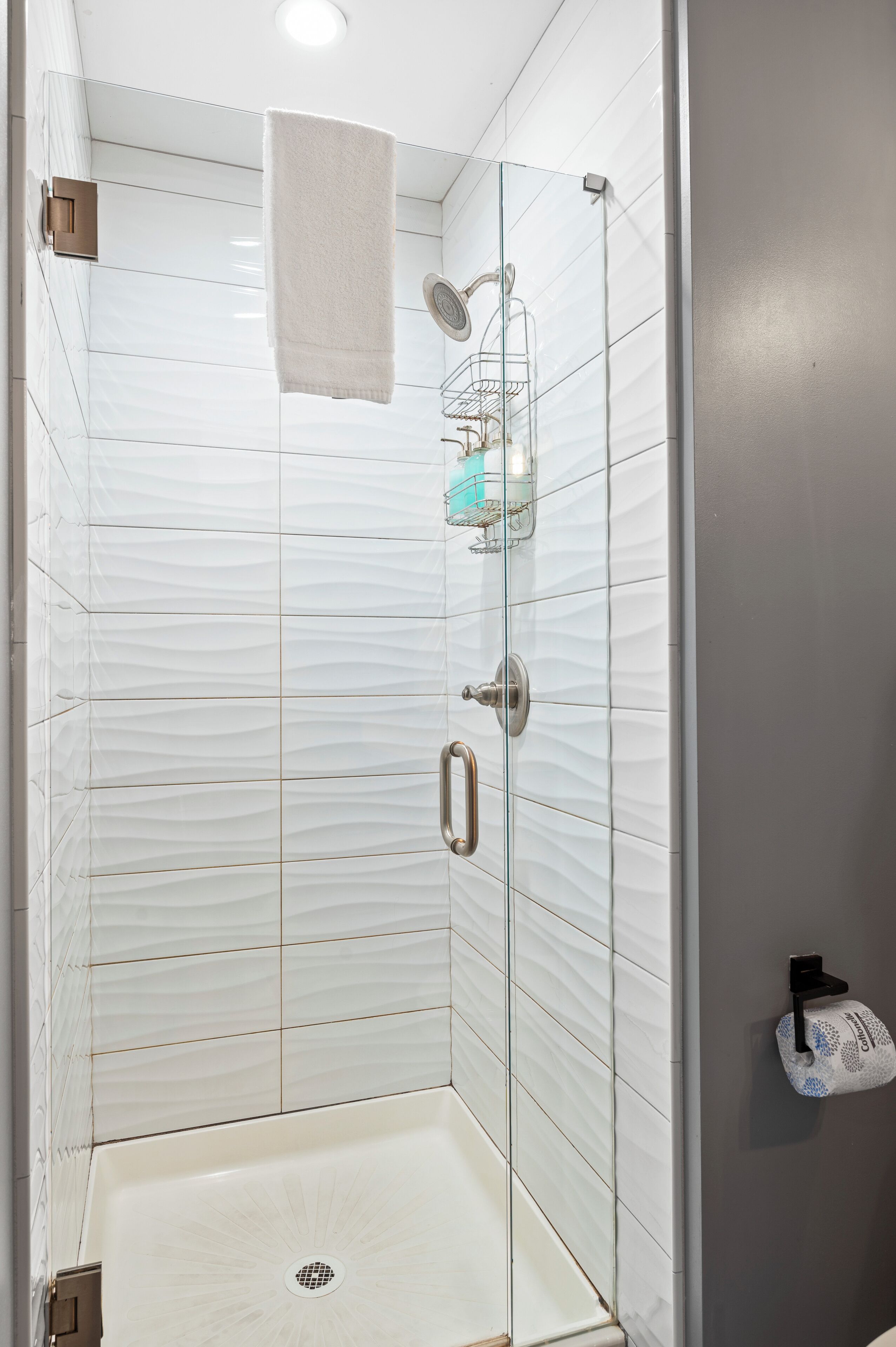 Una ducha a nivel con puerta de vidrio, azulejos blancos con textura, una toalla colgada en la puerta, un jabonera y un sujetapapeles en la pared.
